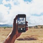 Hand holding a smartphone up against a rural landscape under a cloudy sky