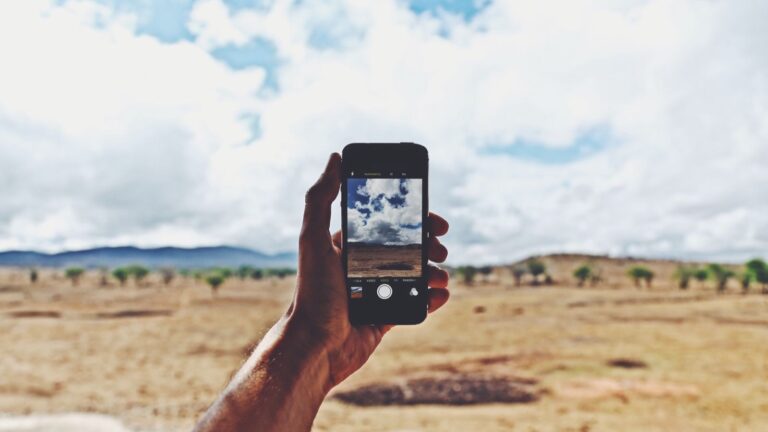 Hand holding a smartphone up against a rural landscape under a cloudy sky