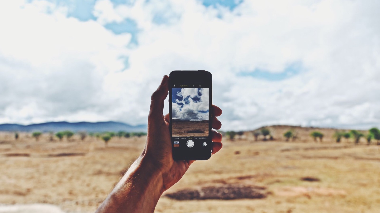 Hand holding a smartphone up against a rural landscape under a cloudy sky