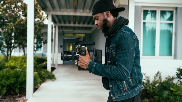 A videographer in a denim jacket and cap films outdoors with a professional camera rig, standing on a covered walkway