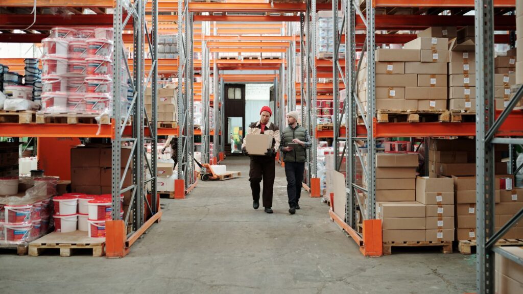 Two warehouse workers carrying a box between shelves stacked with cartons on pallets