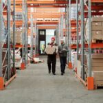 Two warehouse workers carrying a box between shelves stacked with cartons on pallets