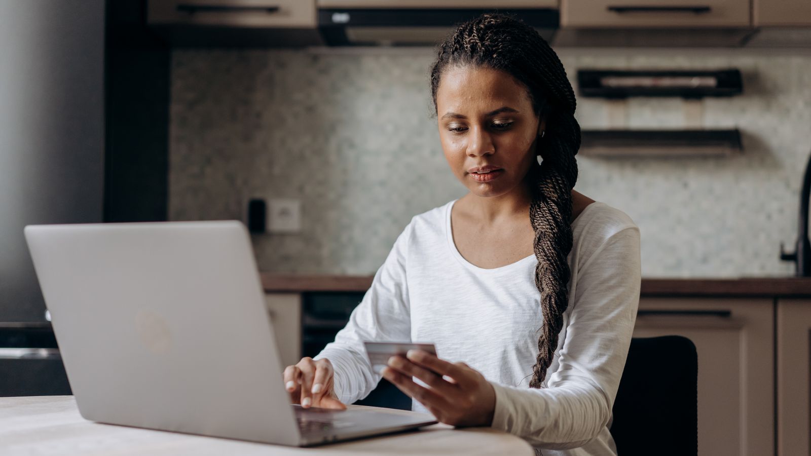 Woman holding a bank card while typing on a laptop at a kitchen table, making an online purchase