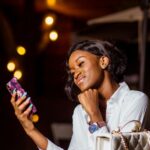 Woman smiling at her smartphone at an outdoor table in the evening, string lights behind her
