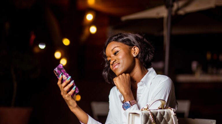 Woman smiling at her smartphone at an outdoor table in the evening, string lights behind her