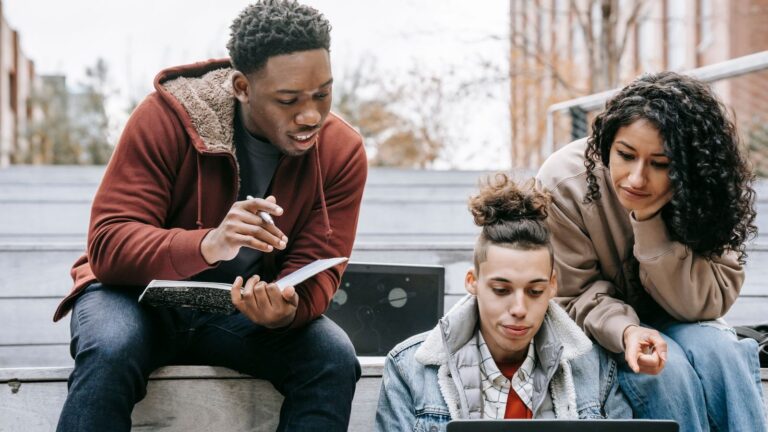 Three students sit on steps on a campus, working together over a laptop and notebook