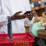 A shopper holds a smartphone and a red grocery basket while shopping in a store.
