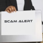 Hand holding a scam alert warning sign next to a laptop