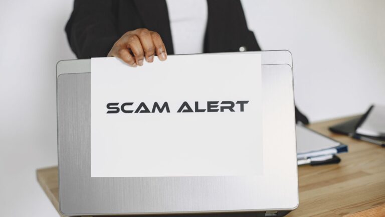 Hand holding a scam alert warning sign next to a laptop